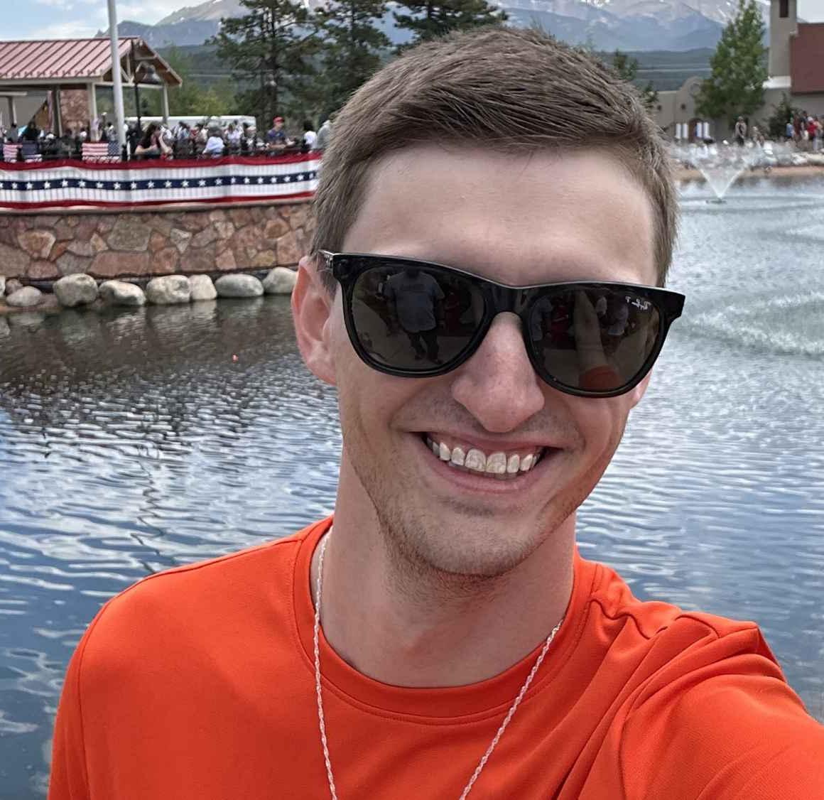 Selfie photo of Logan Ruths, smiling, wearing sunglasses in front of a pond with a flag pole on the other side of the water
